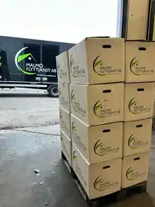 En hög med flyttkartonger märkta Malmö Flyttjänst AB bredvid en flyttbil med varumärket.