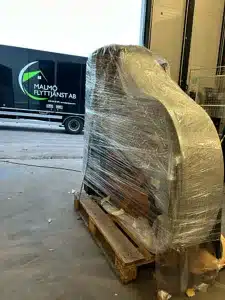 Ett stort piano inslaget i plast ligger på en pall nära en lastbil märkt Malmö Flyttjänst AB.