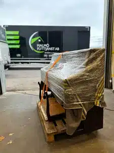 Svart piano inslaget i plast ligger på en pall nära en lastbil märkt Malmö Flyttjänst AB.